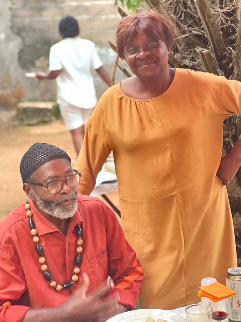 A woman in a mustard yellow dress standing beside a seated man with a beard wearing glasses and a red shirt, both smiling outdoors with a blurred background of a person walking away and a tree.