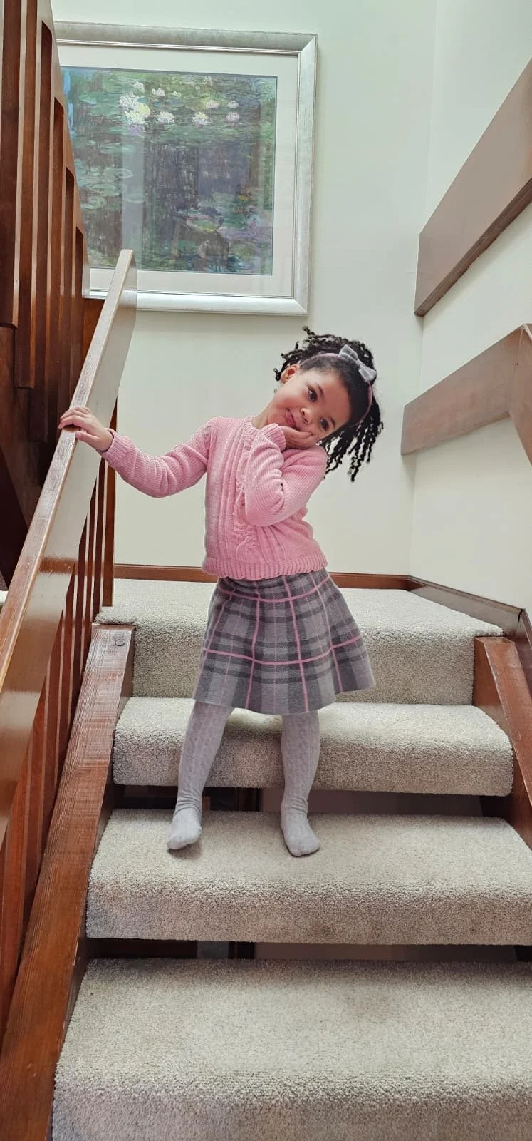 A young girl with braided hair and a bow, wearing a pink sweater, gray plaid skirt, and gray tights, standing on a staircase inside a house, posing with her head tilted and her hand resting on her cheek.