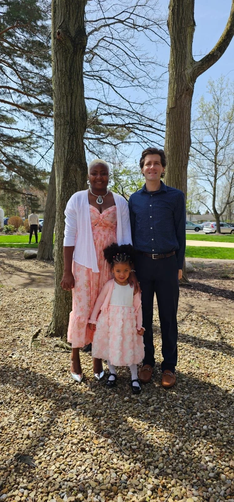 A family of three stands outdoors on a gravel surface. The woman on the left has dark skin and short blonde hair, wearing a peach-colored dress with a white cardigan, and a necklace. The man on the right has light skin and dark hair, dressed in a navy blue shirt and dark pants. A young girl with curly hair, a pink dress with white details, and black shoes stands in front of them, slightly to the right. Behind them are large trees, some with sparse leaves, and a green lawn with a few other people and parked cars in the background.