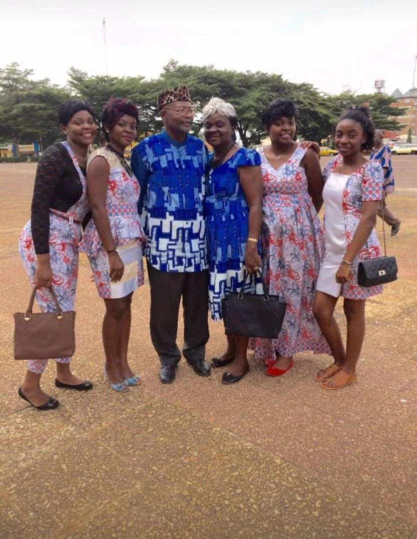 A group of six people, five women and one man, standing outdoors on a dirt area with trees and buildings in the background. They are dressed in coordinated traditional African attire, with most of the women wearing patterned dresses and the man wearing a blue patterned shirt and a traditional hat. All are smiling and posing for the photo.