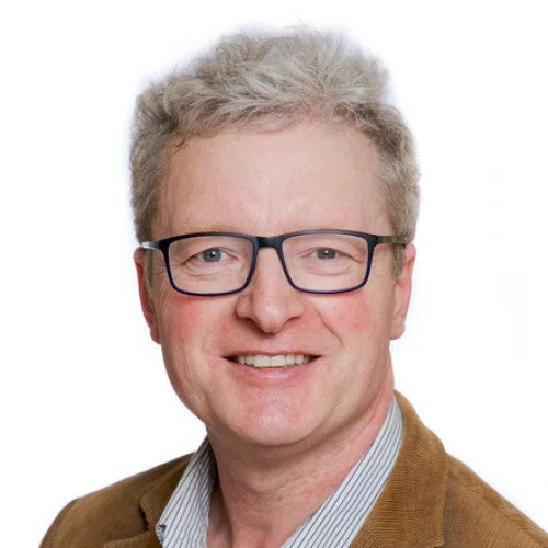 Portrait of a man with curly blond hair, glasses, and a light skin tone, smiling, wearing a brown jacket and a light-colored collared shirt, against a white background.