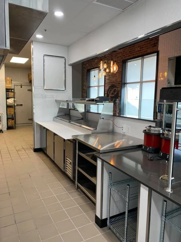 Commercial kitchen with stainless steel counters, a shelf with a food warmer, and kitchen appliances. Two large windows with brick wall backdrop and hanging light bulbs. Storage shelves, tiled floor, and ceiling vents visible.