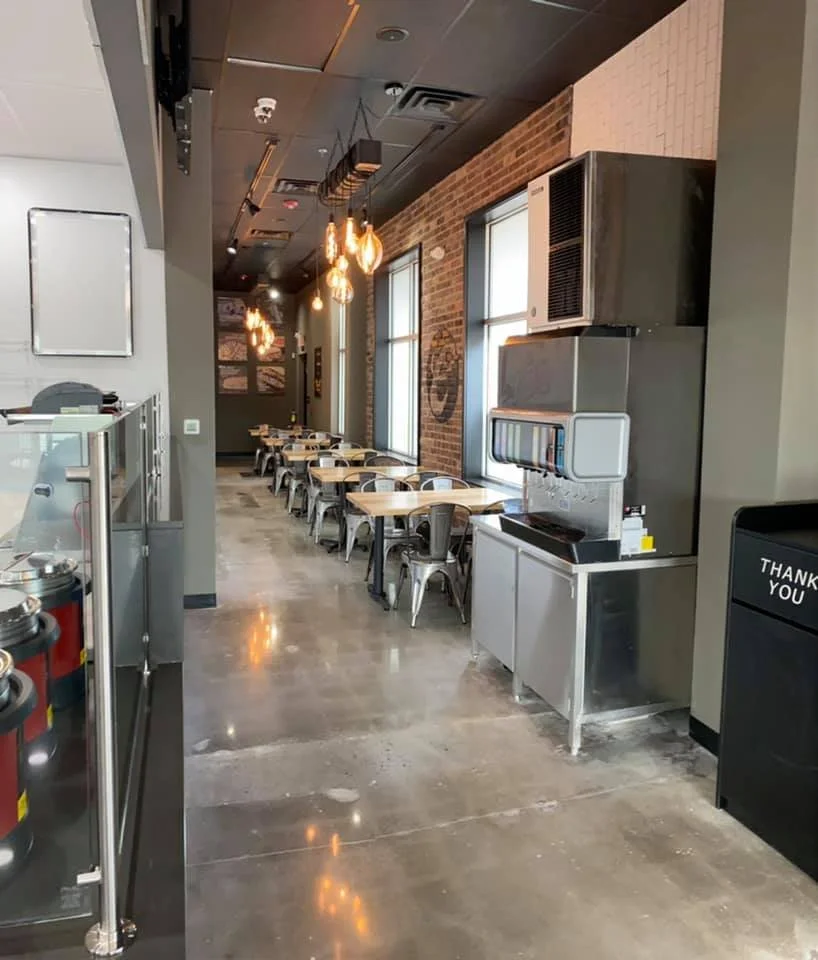 Empty dining tables and chairs along a brick wall with large windows in a modern restaurant.