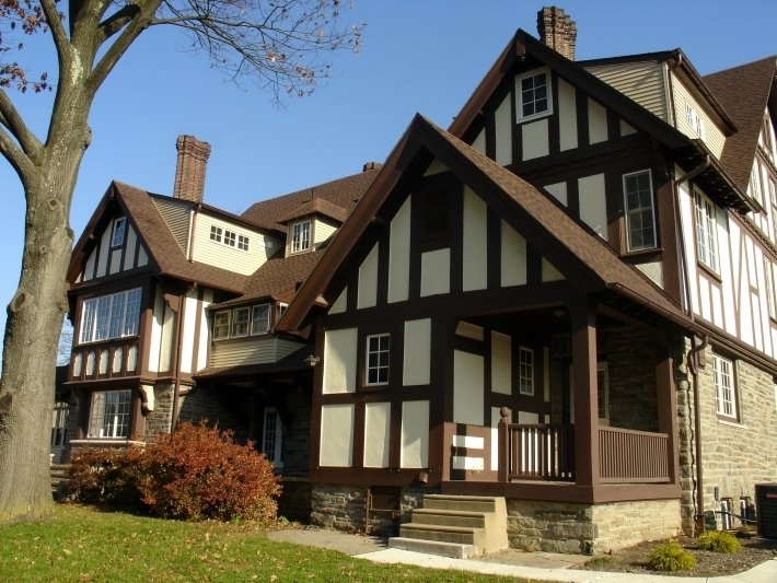 A large, Tudor-style house with a steeply pitched roof, half-timbering, and multiple chimneys, situated in a residential neighborhood with a large tree and landscaping in the foreground.