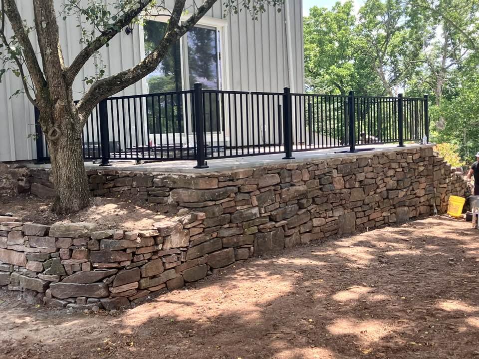A landscaped backyard features a stone retaining wall supporting a concrete deck with a black metal railing, curbside mature trees, and surrounding greenery.