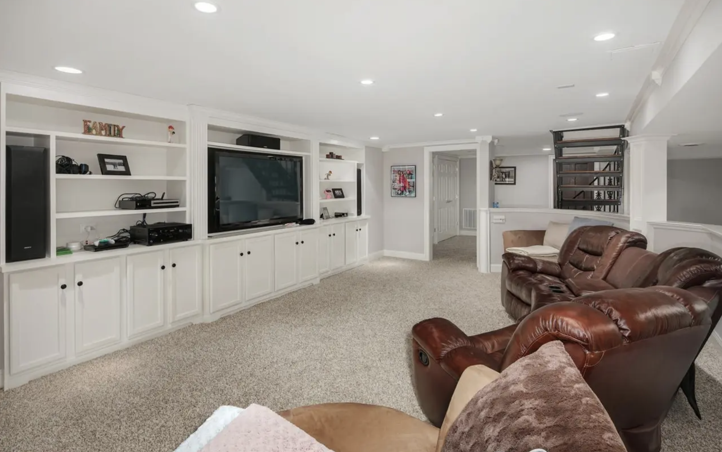 Living room with white built-in shelves and cabinets, large flat-screen TV, brown leather couches, carpeted floor, framed artwork on wall, and a staircase with metal railings in the background.