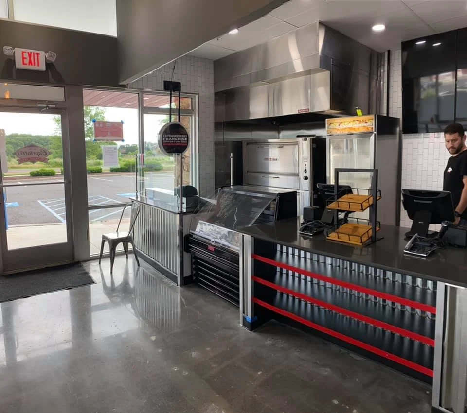 Inside a fast-food restaurant with a counter, stainless steel kitchen equipment, a cash register, and a smiling employee.
