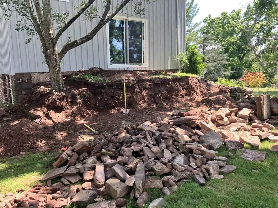 A backyard with a tree, a house with a sliding glass door, and a large pile of rocks and dirt indicating ongoing landscaping or foundation work.