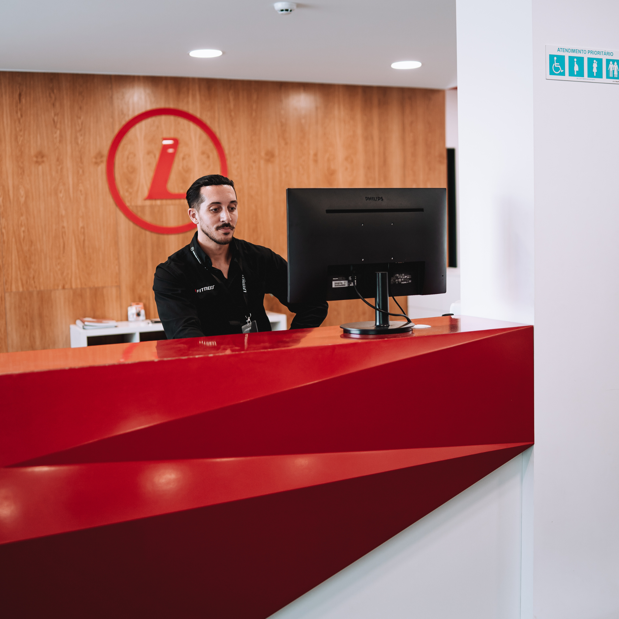 Recepcionista trabalhando na recepção de uma clínica, com fundo de parede de madeira e logo vermelho de uma cruz e 'L' na parede, e um painel de sinais de acessibilidade amparando deficientes e idosos.
