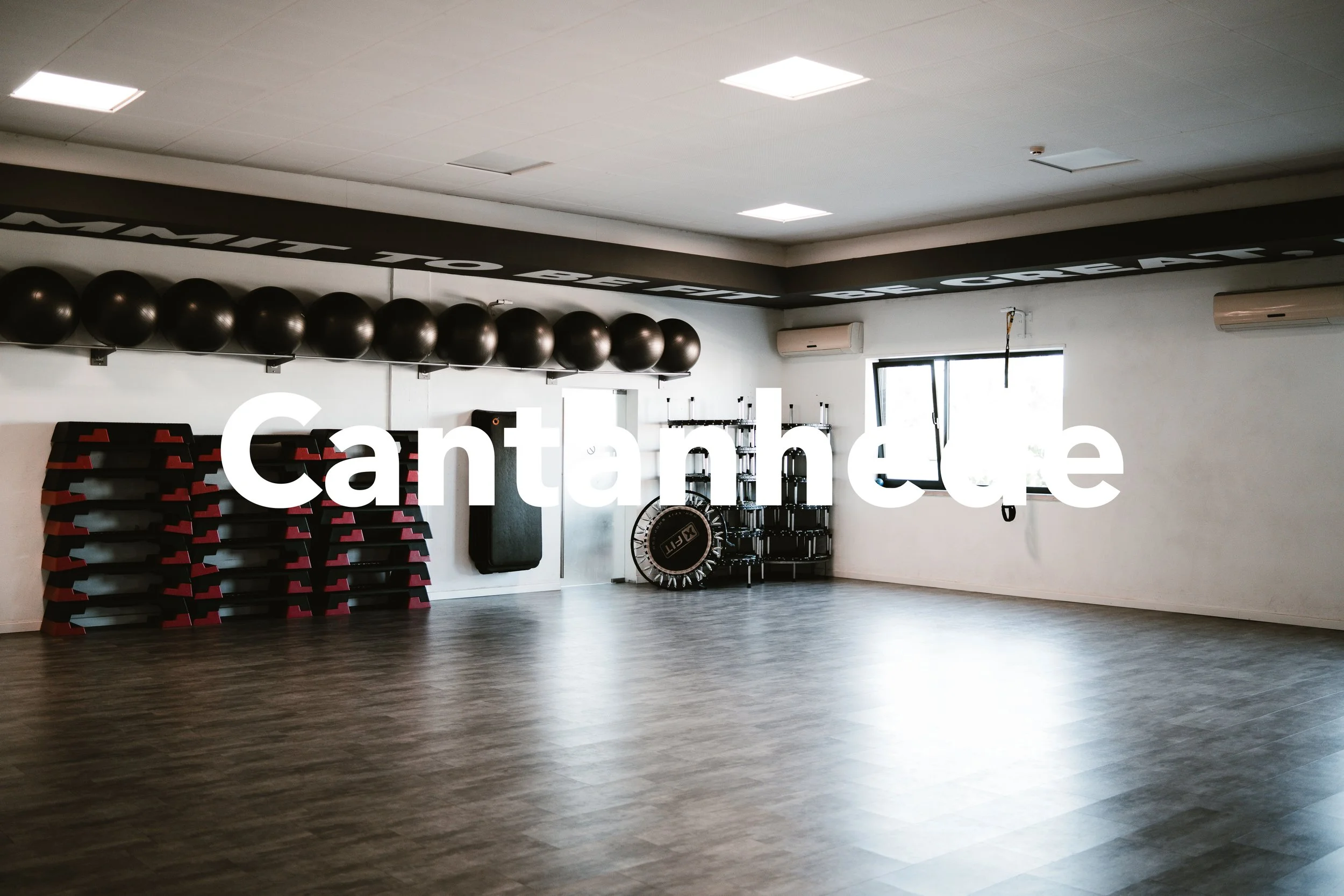 Sala de ginástica vazia com bolas de exercício, plataformas, saco de boxe, trampolim e uma janela, ambiente bem iluminado e limpo.