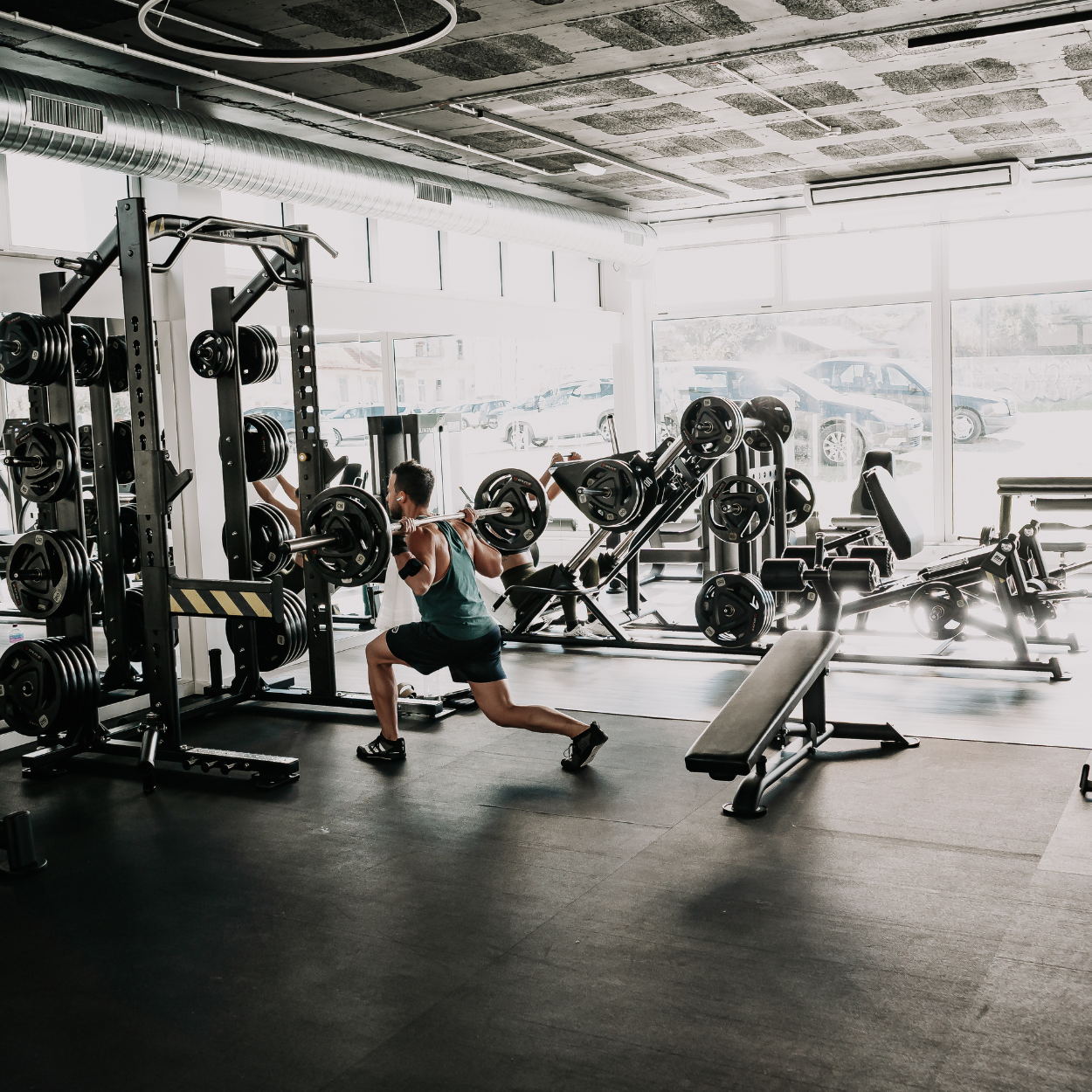 Pessoa fazendo agachamento com barra em academia bem iluminada com muitas máquinas de exercícios.