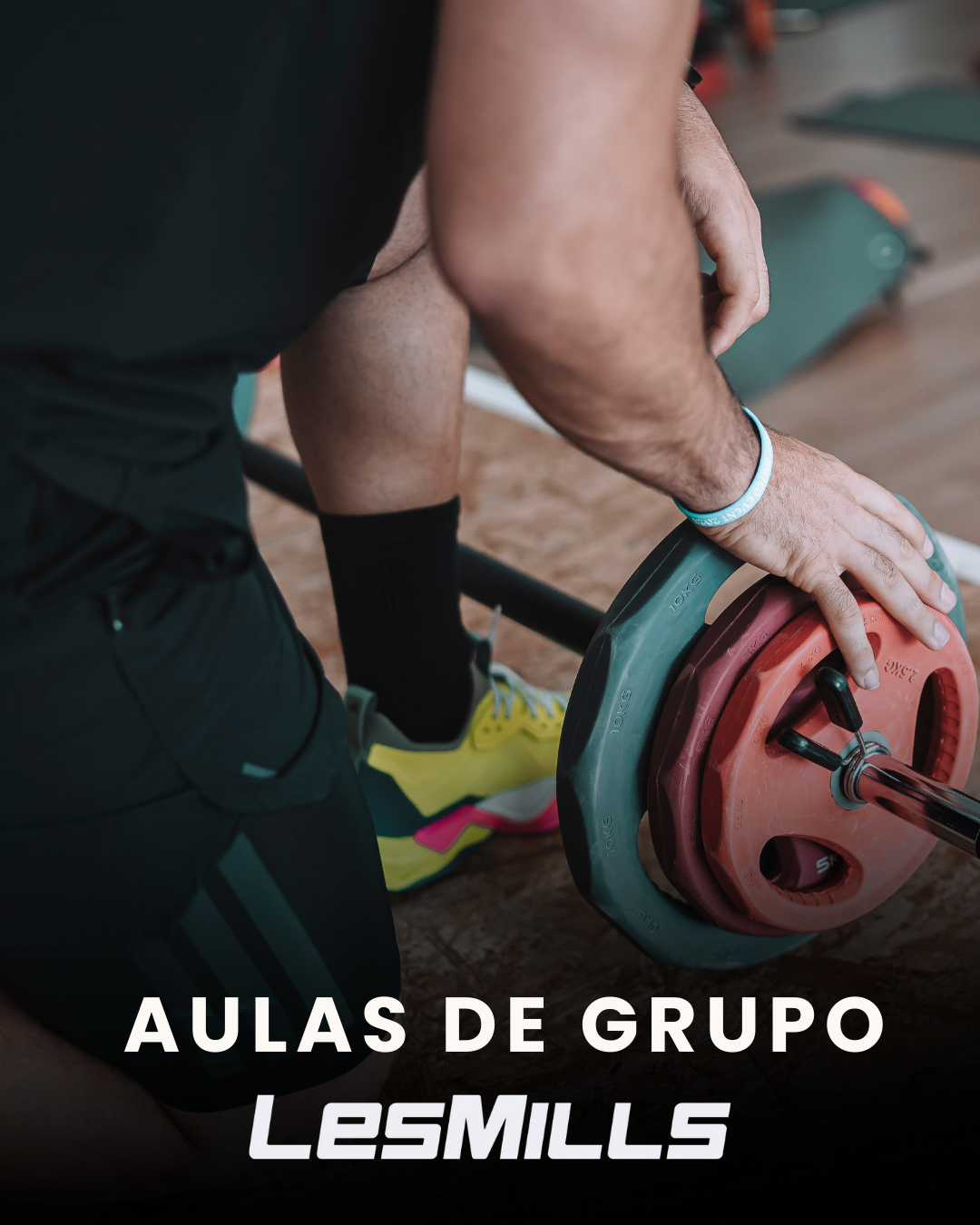 Pessoa se exercitando na academia, aproximada de um haltere com peso, usando roupa confortável e tênis esportivos. Texto na imagem: Aulas de Grupo LesMILLS.