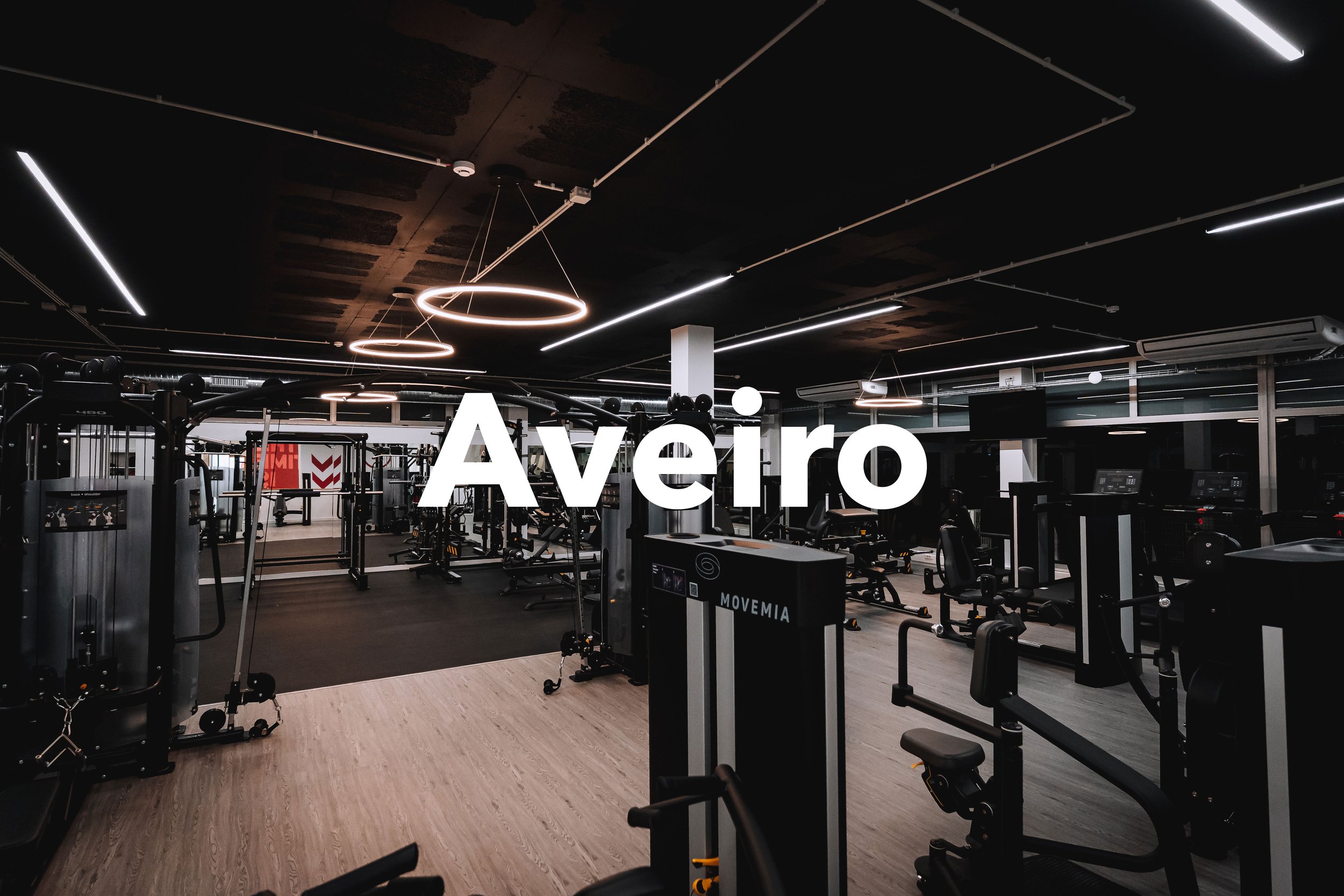 Imagem de uma academia moderna com equipamentos de ginástica, iluminação de LED no teto preto e janelas grandes. A palavra 'Aveiro' está escrita em destaque na imagem.