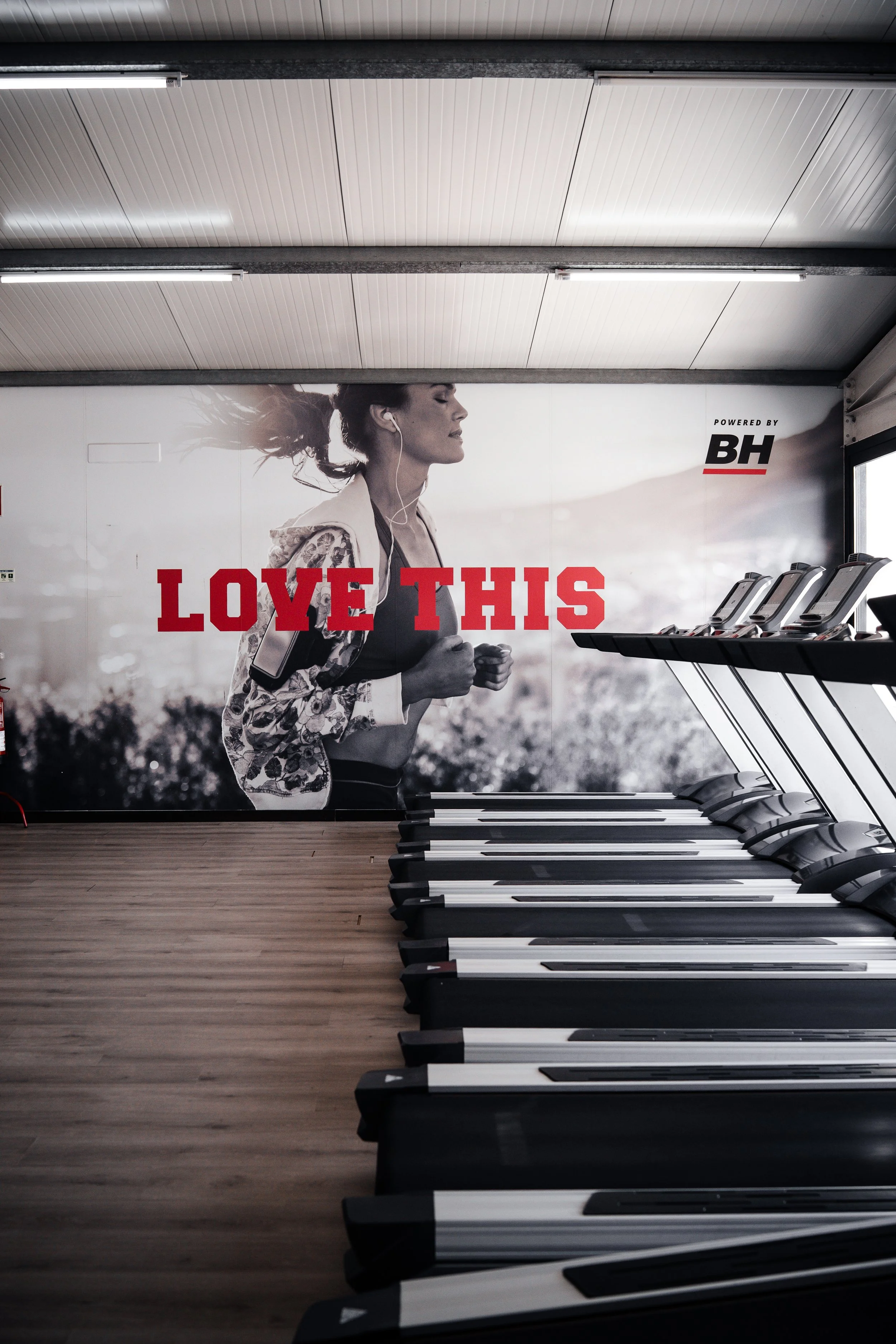 Piso de academia com esteiras de exercício ao lado de uma parede com pôster de uma mulher correndo, com a mensagem 'LOVE THIS' em destaque.