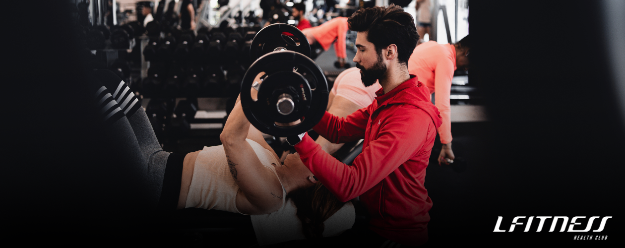 Homem treinando com halteres na academia enquanto uma mulher deitada na esteira faz exercício.
