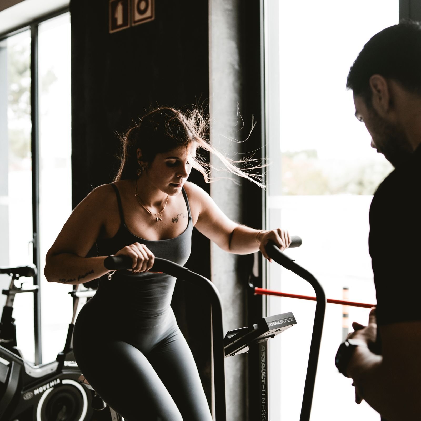 Mulher usando máquina de exercícios, supervisionada por instrutor, em academia com iluminação natural vindo de uma janela.