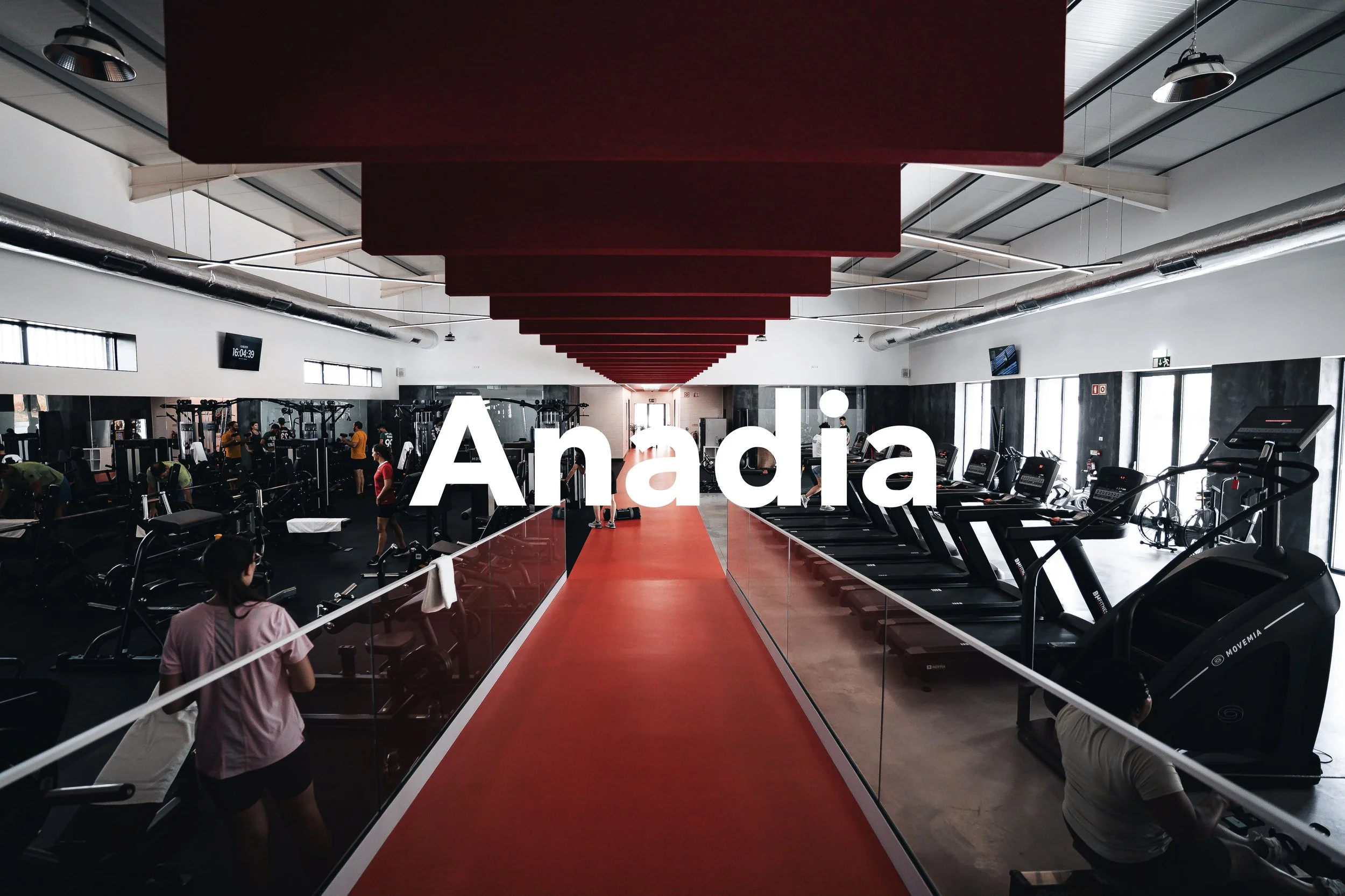 Interior de uma academia de ginástica moderna, com esteiras e equipamentos de musculação, pessoas treinando e um corredor vermelho central.