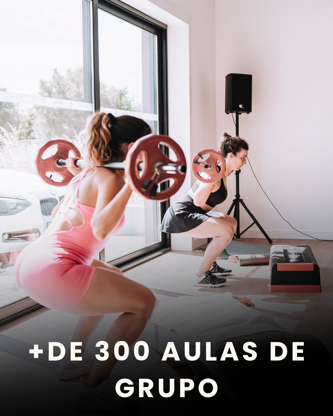 Duas mulheres fazendo agachamentos com barras de peso em uma academia, com uma grande janela ao lado.