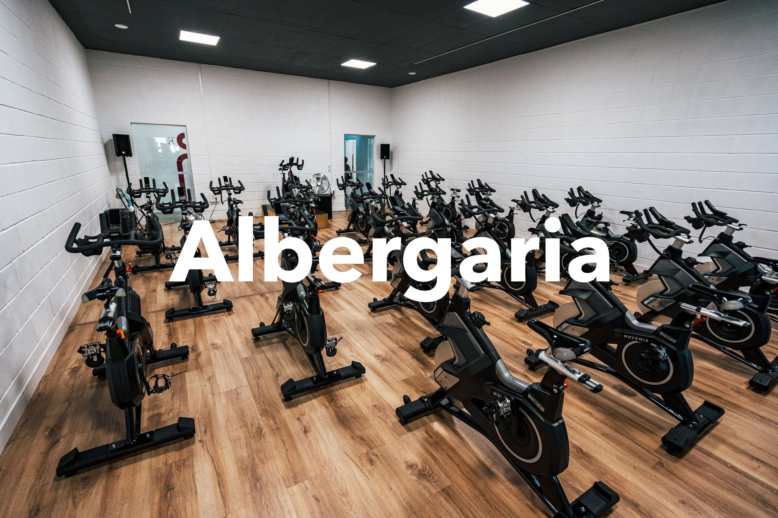 Sala de spinning com várias bicicletas ergométricas pretas e parede branca, com o texto 'Albergaria' overlay.