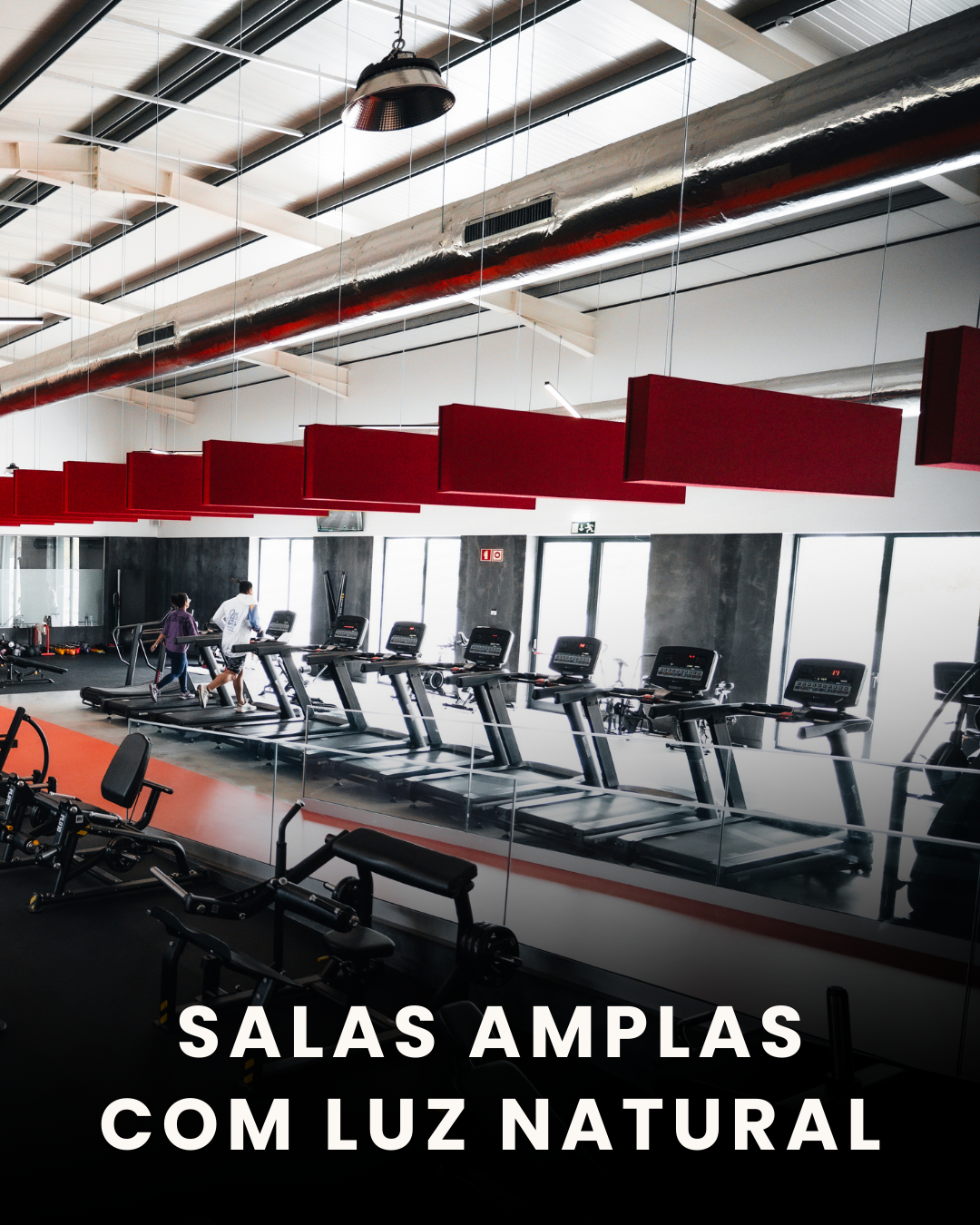 Sala de ginásio ampla com luz natural, plataformas de corrida, equipamentos de musculação e pessoas treinando.