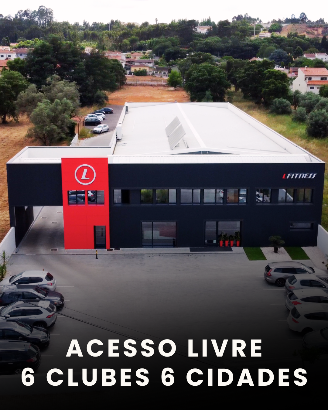 Prédio moderno de academia de ginástica com fachada preta, porta de entrada, janelas grandes e um símbolo circular vermelho com a letra 'L' na parte superior. Carros estacionados na frente e sinais de acesso livre na mensagem escrita na imagem.