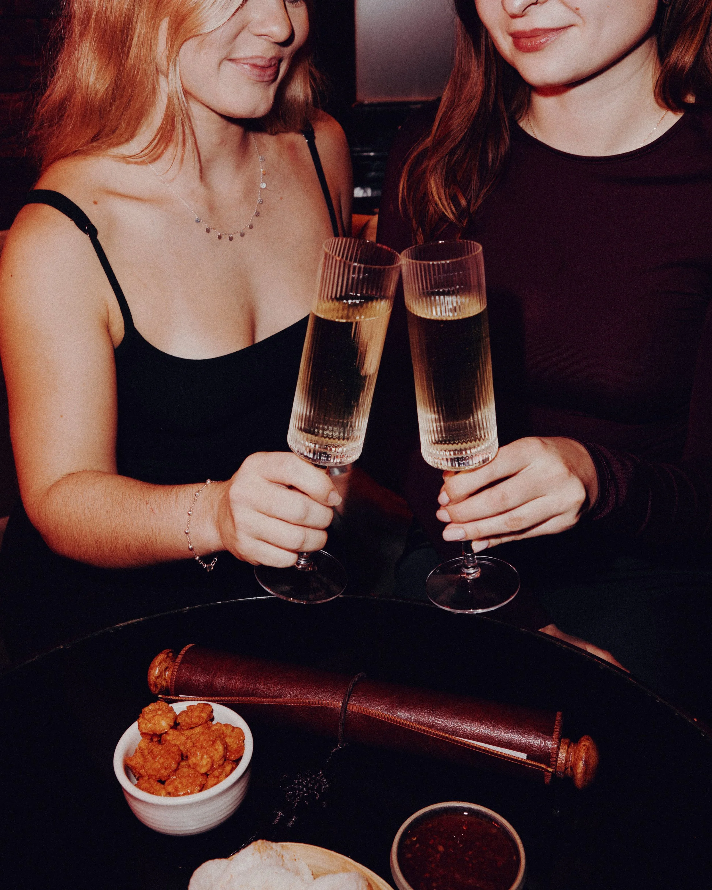 Two women holding champagne glasses in a toast, with a small bowl of fried food and dipping sauce on a table in front of them.