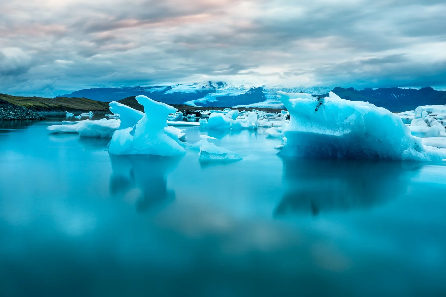 Glacial icebergs floating in calm arctic water in Iceland with distant mountains and a cloudy sky in the background.