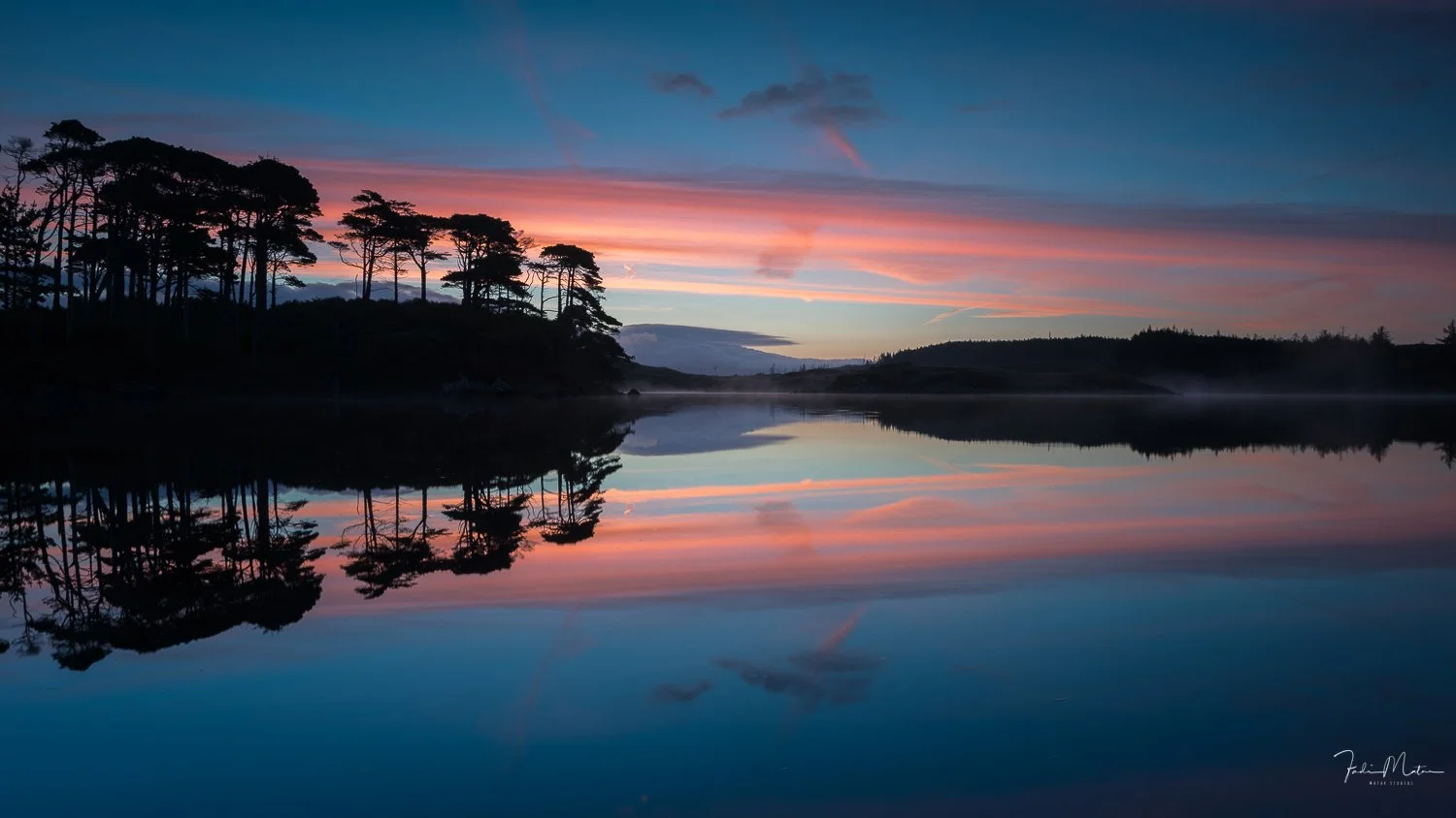 Calm lake reflecting trees and colorful sunrise sky with pink, orange, and blue hues.  This is one of the many beautiful scenes I experienced in Ireland