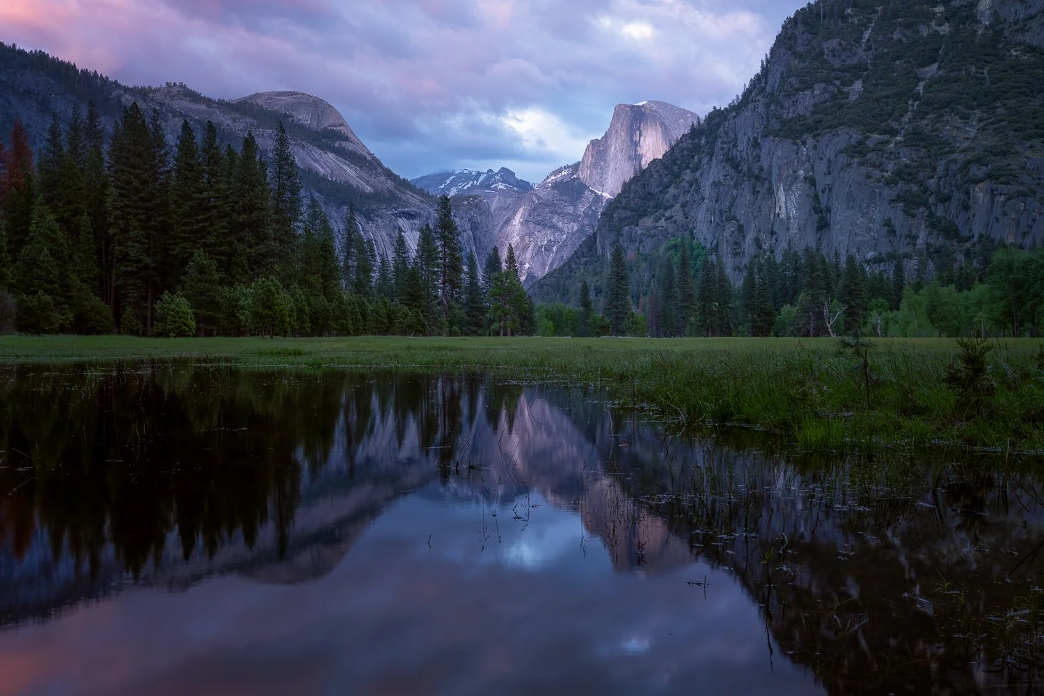 Scenic view of a mountain landscape with a reflective body of water in the foreground, pine trees, and towering granite cliffs under a cloudy sky.