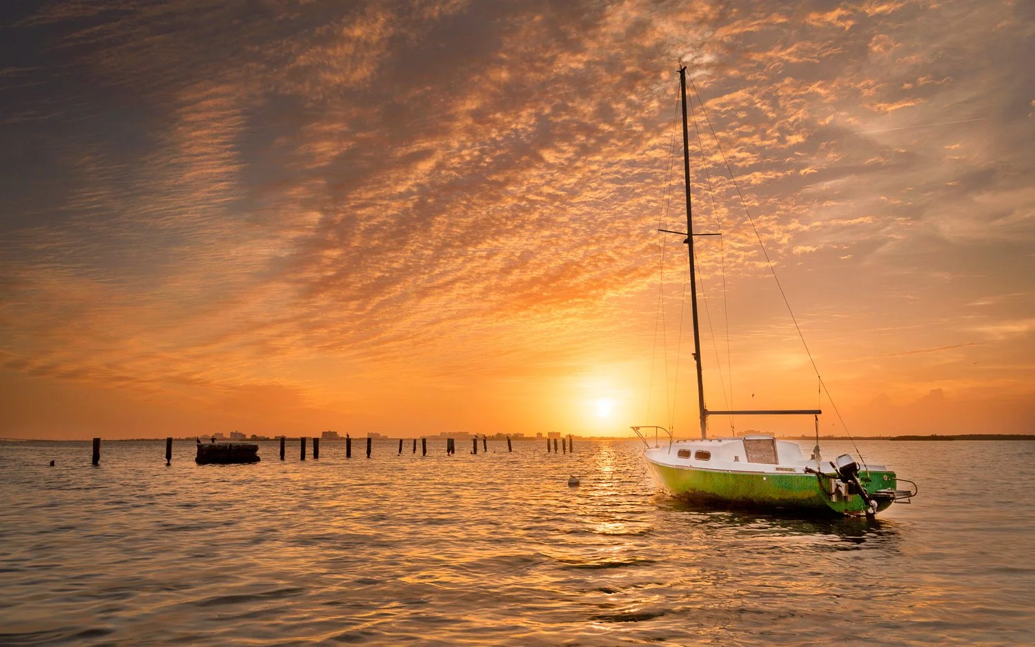 A sailboat floating on calm waters in Tampa Bay during a sunset with a colorful sky and a bright sun near the horizon.