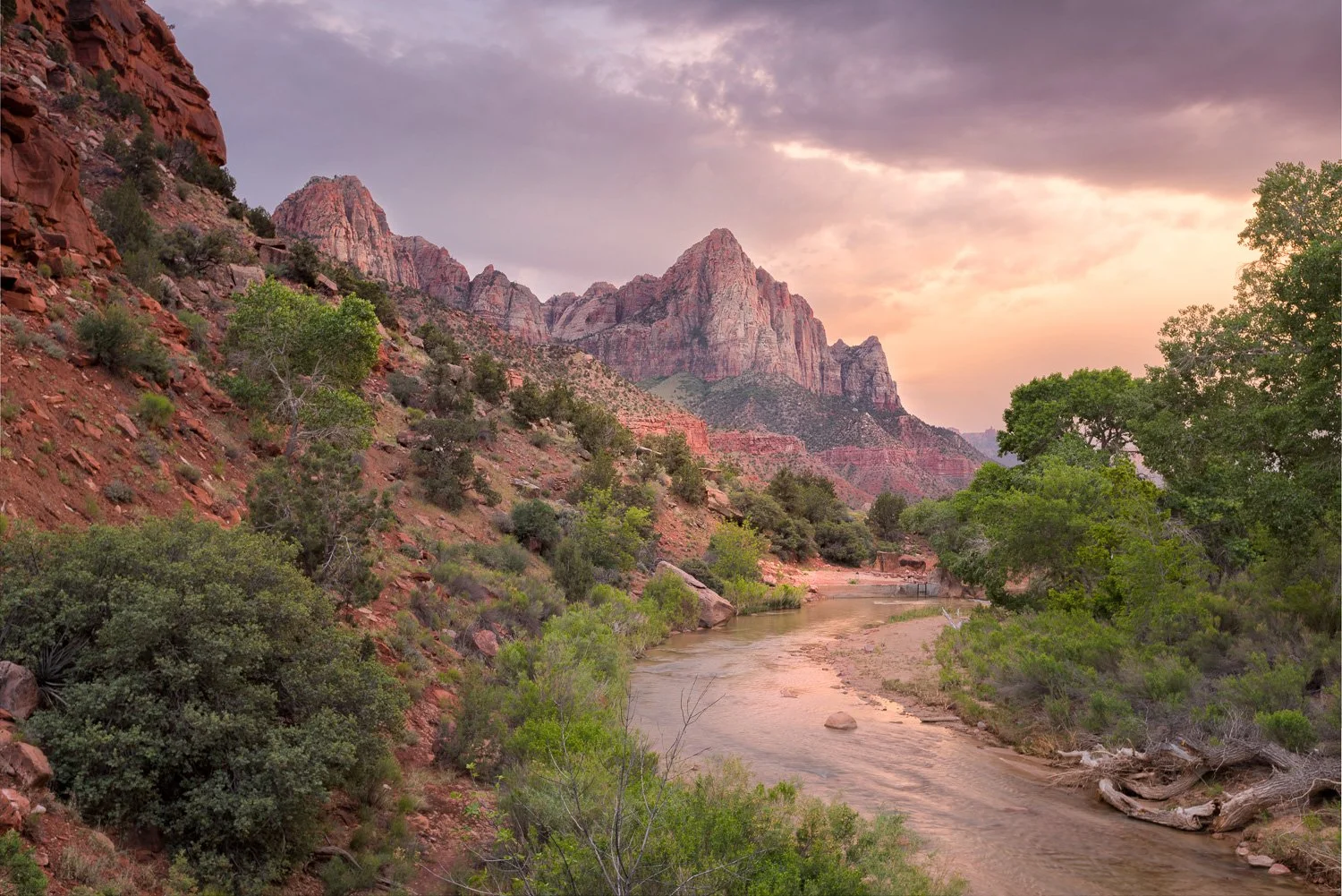 A landscape of Zion National Park featuring red rock mountains, a river flowing through green trees, and a colorful sky at sunset.