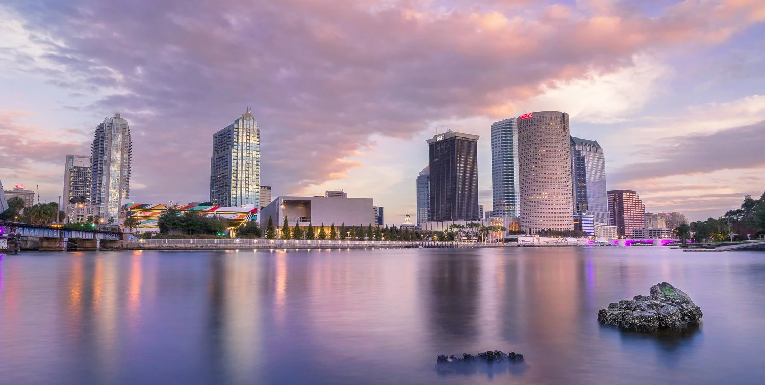 Tampa City skyline at dawn over a calm Hillsborough River with colorful clouds in the sky and rocks in the water. I took this shot from the University of Tampa campus