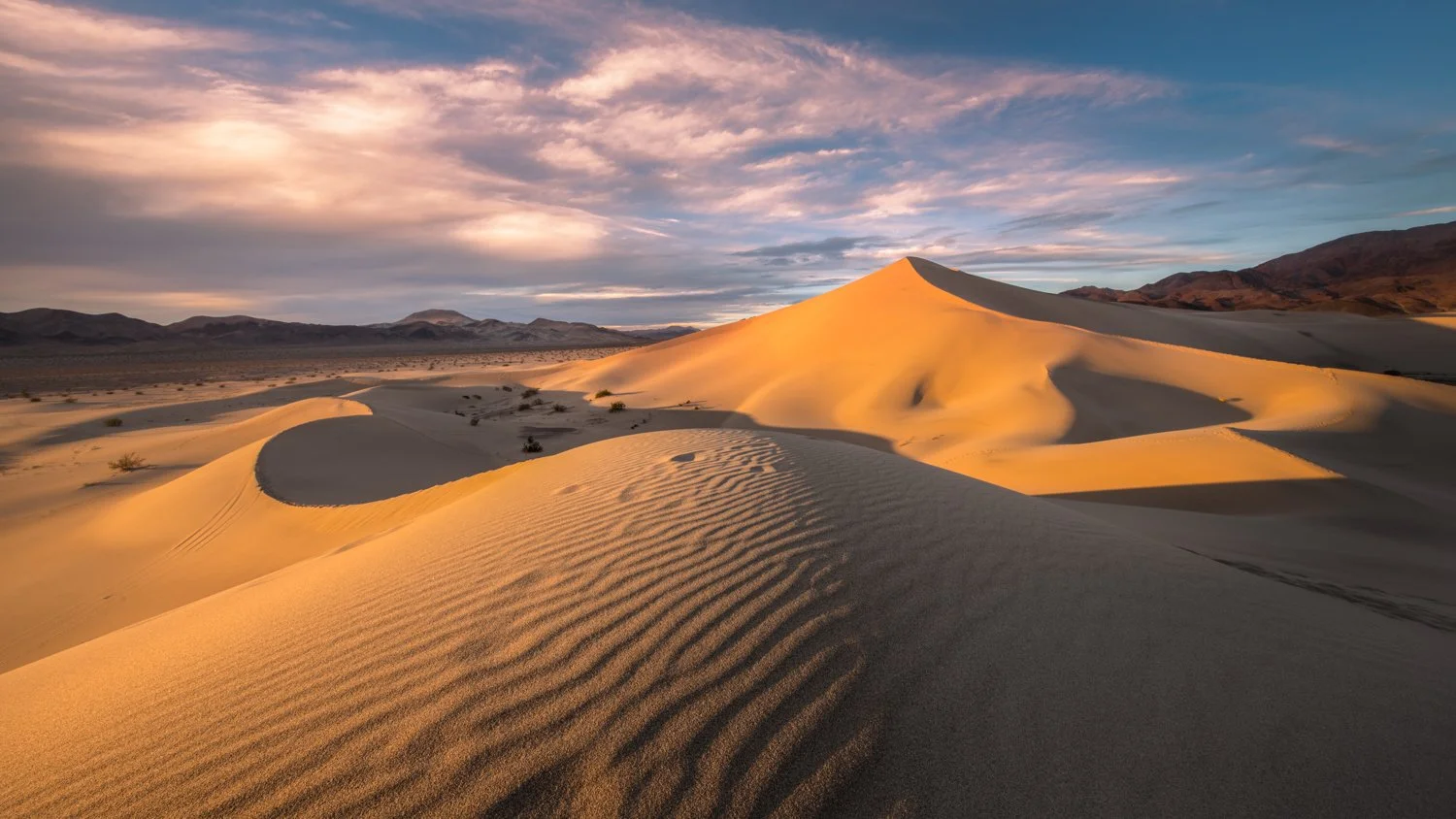 Sand dunes in Death Valley during sunset with mountains in the background and a partly cloudy sky.