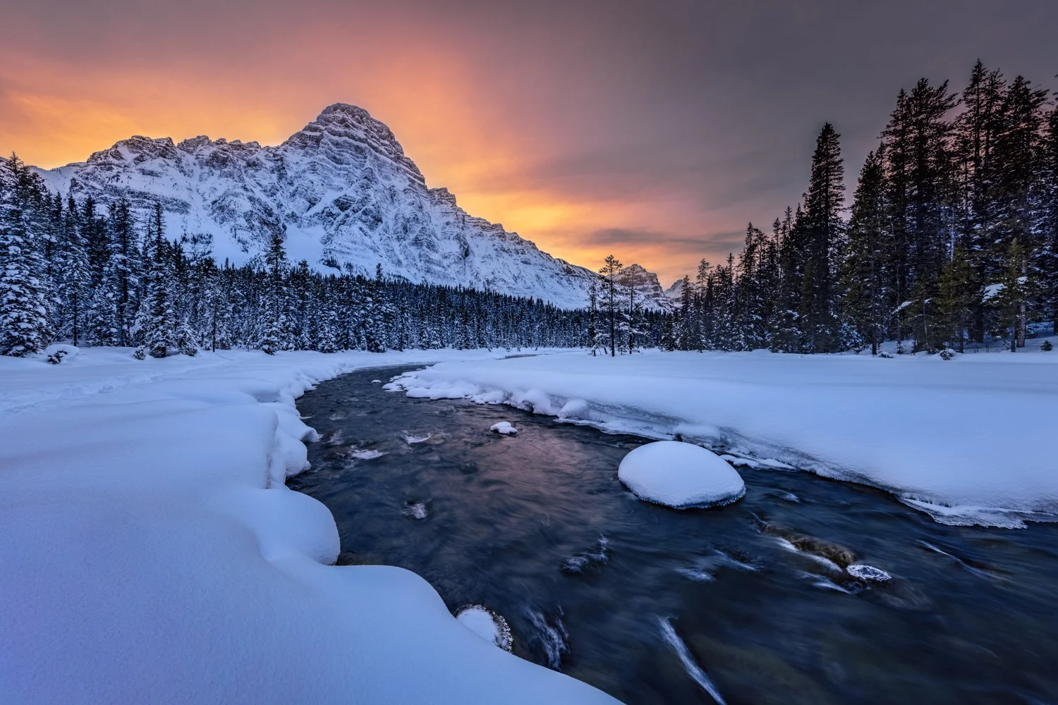 Snow-covered landscape featuring a flowing river, dense evergreen trees, and a mountain range at sunset with a colorful sky. Shot taken in the Canadian Rockies