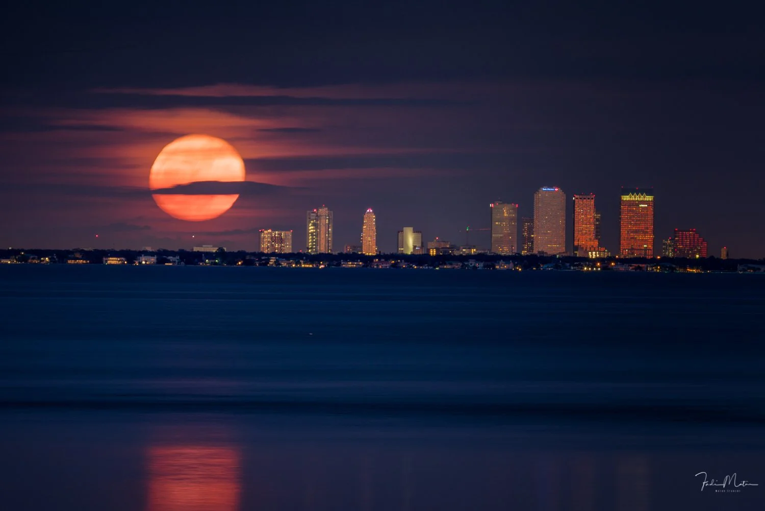 Nighttime city skyline with tall buildings and illuminated windows, moon partially obscured by clouds, reflected on calm water in the foreground.