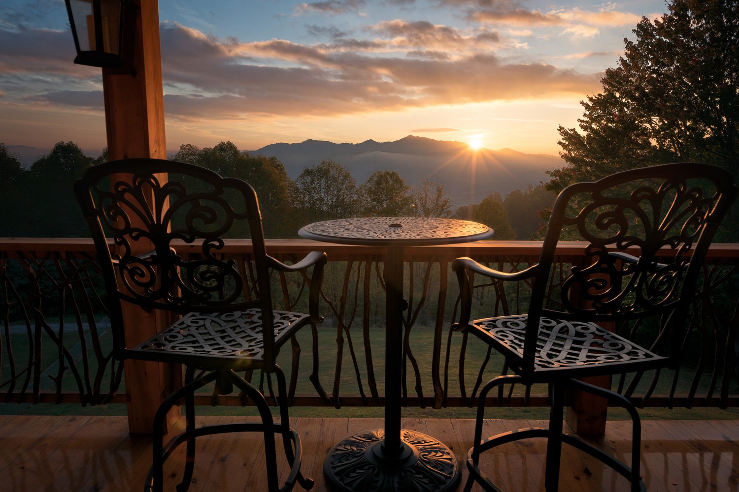 Sunset view from a wooden deck with two ornate metal chairs and a round table, overlooking mountains and trees.