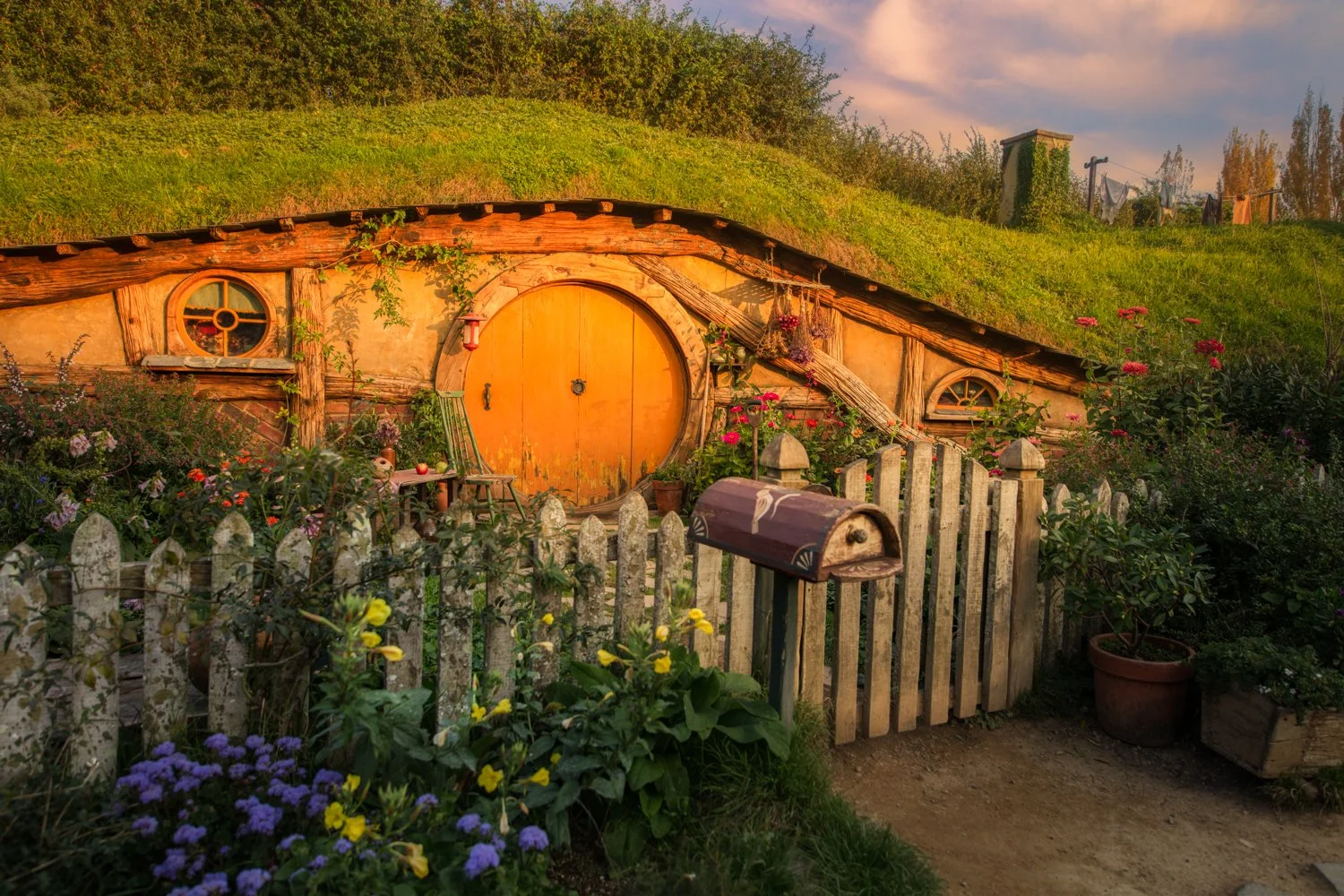 A Hobbit-style house with a round, orange door set into a hillside, surrounded by a colorful garden with flowers and plants, a wooden fence, and a mailbox near the entrance, during sunset.  This is one of the Hobbit houses in the Hobbiton movies in N