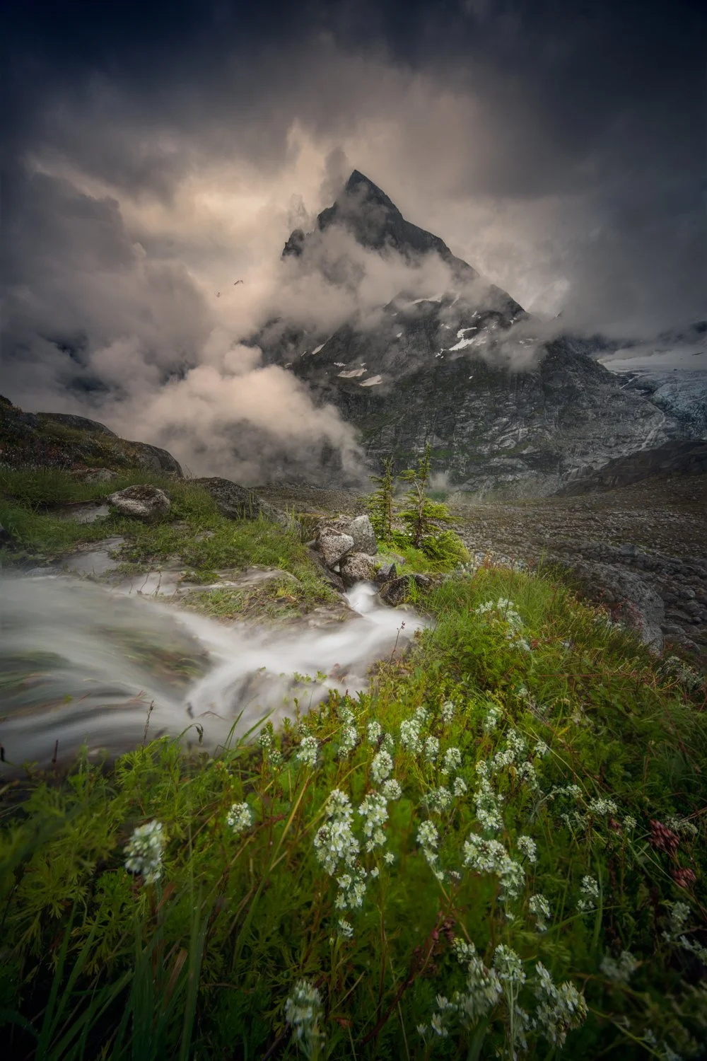 A dramatic mountain scene with a rugged, snow-capped peak shrouded in dark clouds, a rushing stream with white water flowing over rocks, lush green grass, and wildflowers in the foreground.