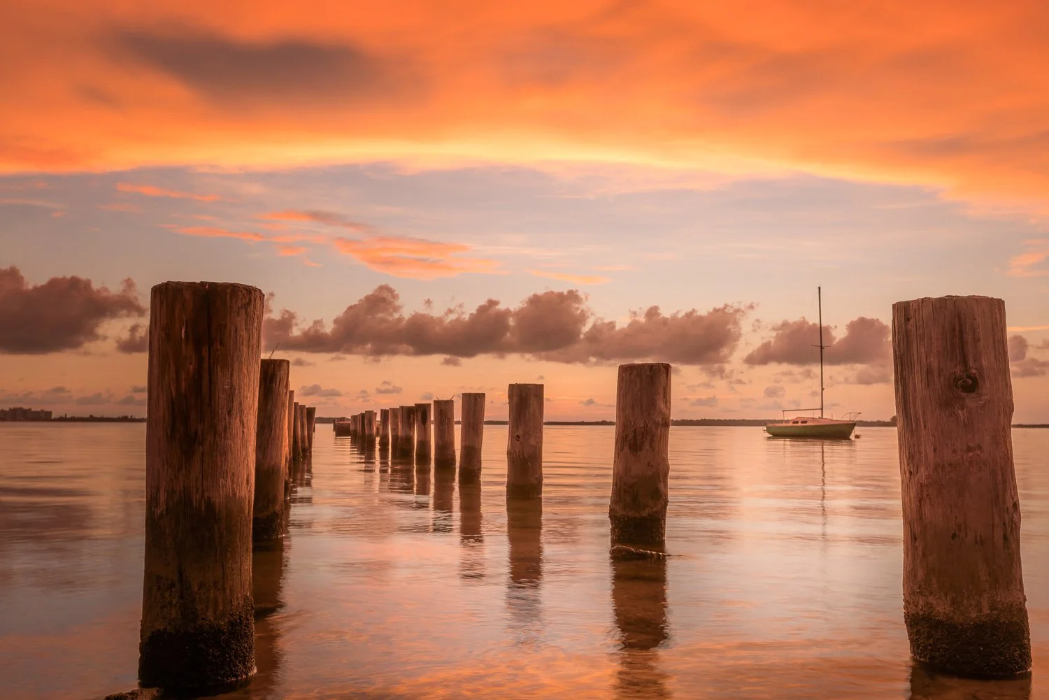 A calm body of water at sunset with wooden pilings in the foreground and a sailboat in the distance, under a colorful sky with orange and pink clouds.