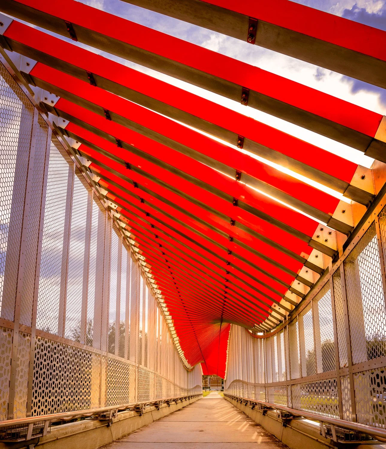 A pedestrian bridge with a red, curved overhead canopy and metal side panels, illuminated by sunlight, leading towards a path with trees and sky visible in the background.