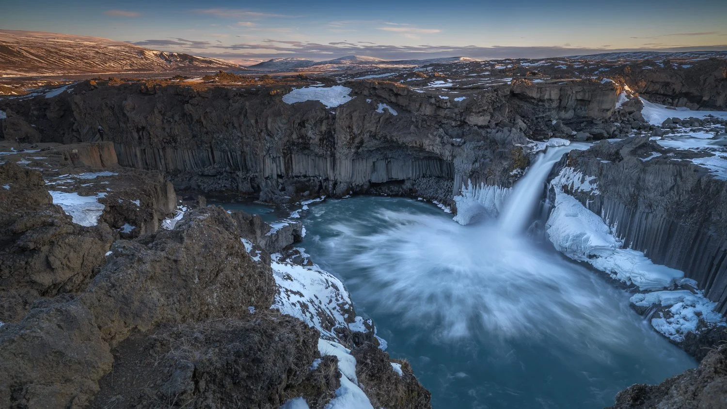 A waterfall flowing into a river in a snowy, rocky canyon with hills and a partly cloudy sky in the background.  This shot is taken in Iceland