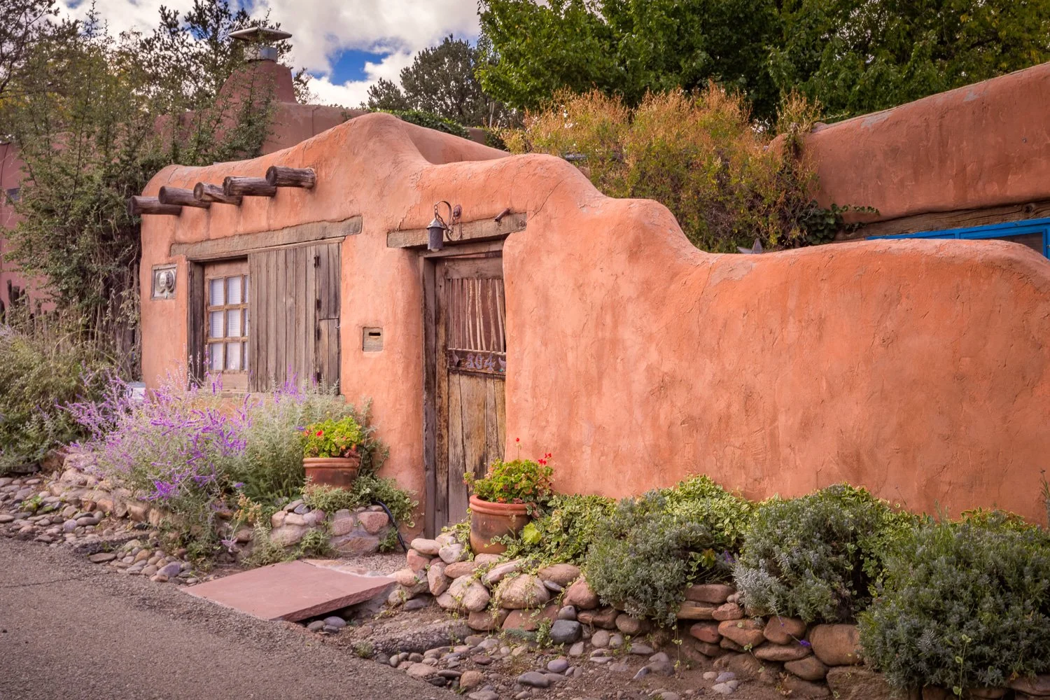 An adobe-style building with a rounded, textured exterior, a wooden door, and a small window surrounded by potted plants and rocks. Greenery and trees are visible in the background under a partly cloudy sky.