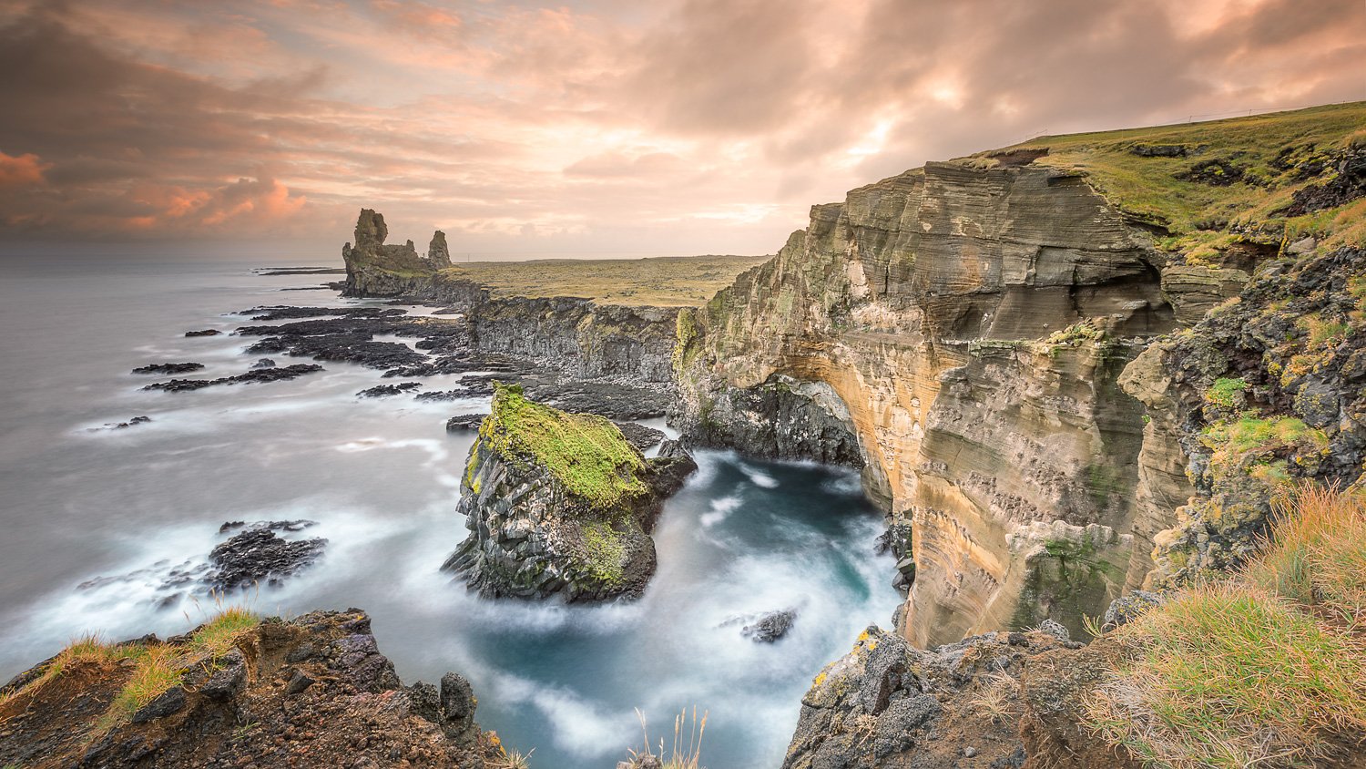 Cliffside landscape in Iceland, with rugged rocks green moss, and ocean waves crashing below, under a cloudy sky at sunset.