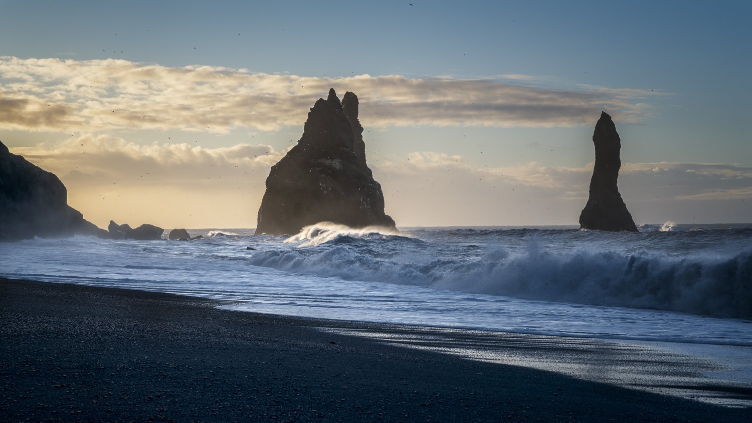 Ocean waves crash on a black sandy beach in Iceland with large rock formations in the water and a partly cloudy sky at sunset.