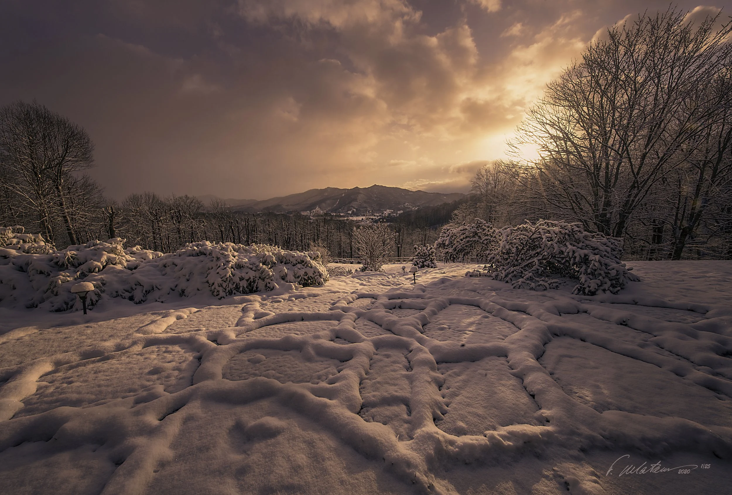 Snow-covered landscape with trees, bushes, and a mountain range in the distance during a sunset or sunrise.  I took this shot in front of our mountain home in Waynesville, North Carolina on Christmas morning.  That year we had true white Christmas Ev