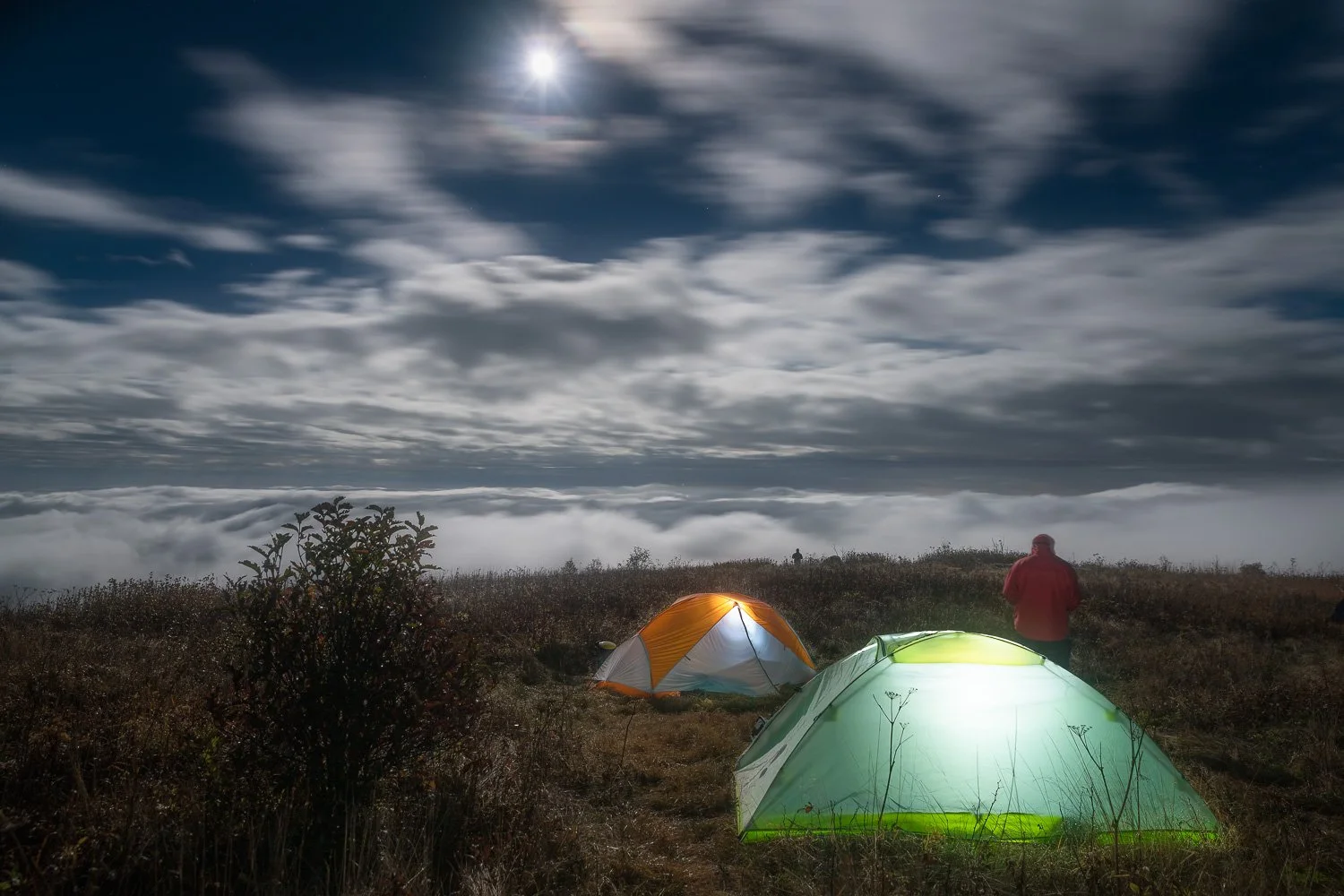 Nighttime camping scene with two tents, one orange and white, and one green, on a grassy hill under a cloudy sky and bright moon, with my buddy, Chris as he poses in a red jacket trying to stand still during the long shutter speed shot near the tents