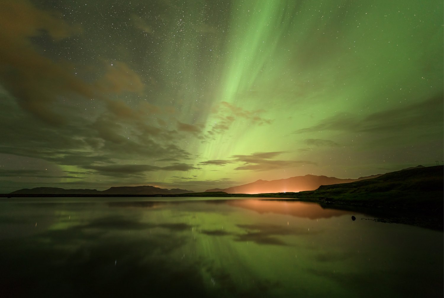 Night sky illuminated by the green Northern Lights over a calm lake with mountain silhouettes in the distance.