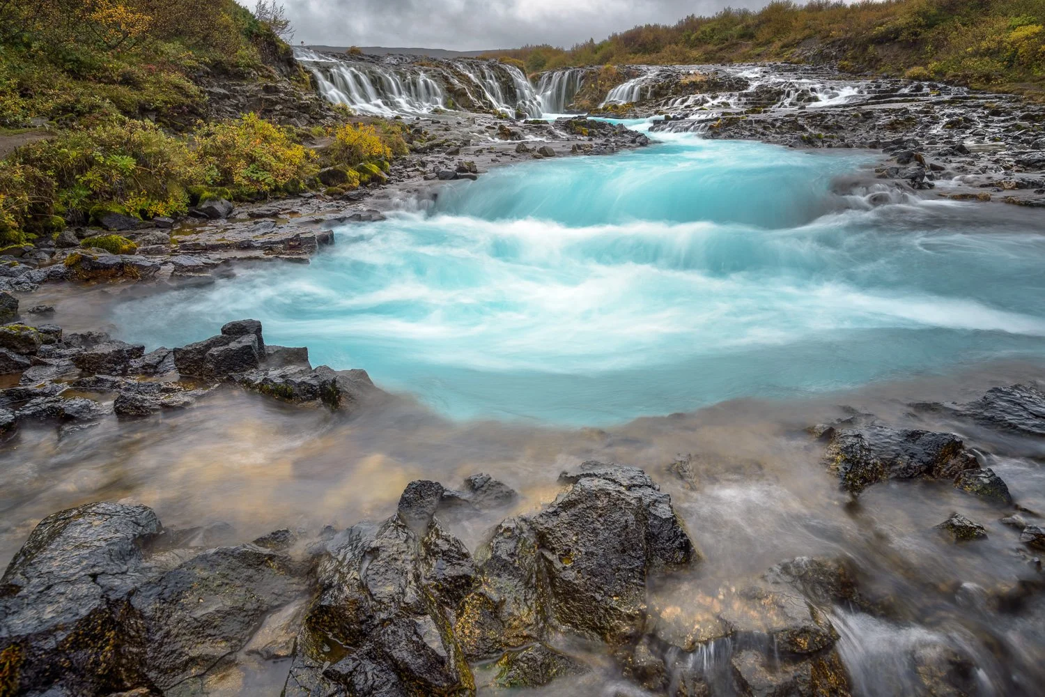 A waterfall flowing into a bright blue pool with dark rocks around it, surrounded by greenery and overcast sky.