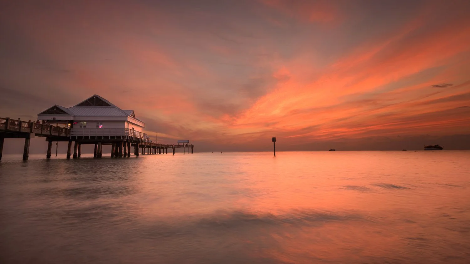 Sunset over a pier extending into calm ocean waters, with a building on stilts, a ship in the distance, and colorful sky with orange and pink clouds.