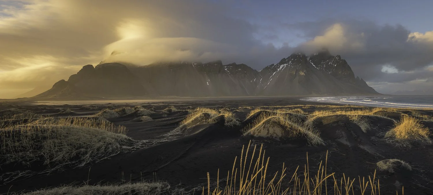 A black sand beach with dunes and tall grass, with mountains in the background under a partly cloudy sky during sunset or sunrise.  This scene is from around Vestrahorn mountain in Iceland